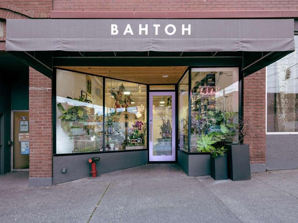 Exterior of BAHTOH salon, a brick storefront with a gray awning reading 'BAHTOH,' large display windows filled with plants and flowers, and a neon sign glowing inside