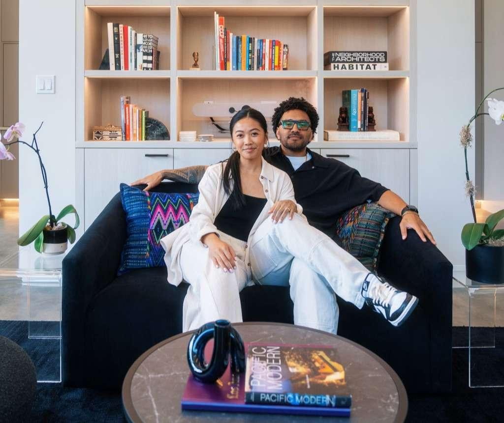 Sonia-Lynn and Sergio Max, founders of La Union Studio, sitting together on a black sofa in front of built-in shelving styled with books and decor, with a coffee table displaying the book 'Pacific Modern