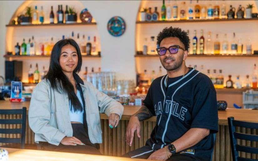 Sonia-Lynn and Sergio Max sitting together at a bar counter, with shelves of liquor bottles illuminated behind them