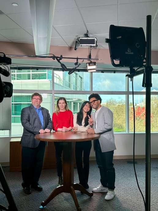 Four people posing together in a TV studio setup with cameras and lighting equipment, standing around a round table during a KOMO News interview
