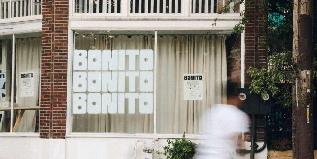 Exterior of Bonito Café y Mercadito storefront with large 'BONITO' text on the windows, brick facade, and a motion-blurred cyclist passing by