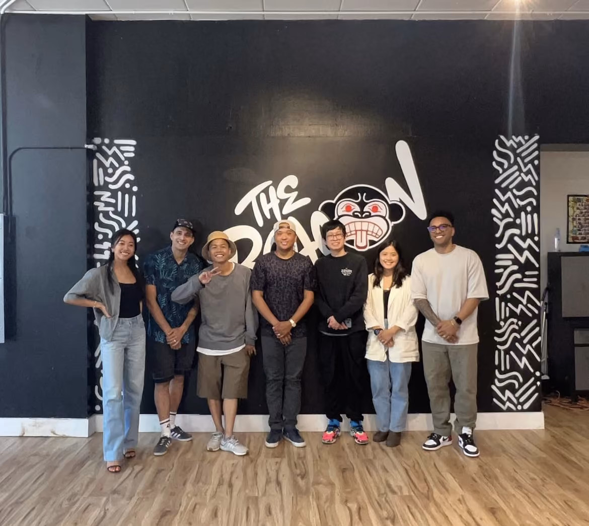 Eight people posing together in front of a black wall with a white graffiti-style mural reading 'The Baboon