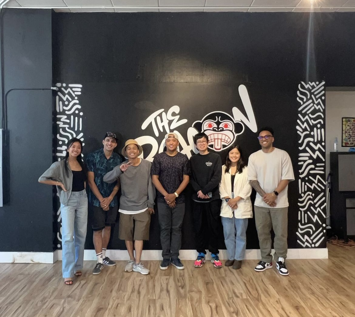 Eight people posing together in front of a black wall with a white graffiti-style mural reading 'The Baboon