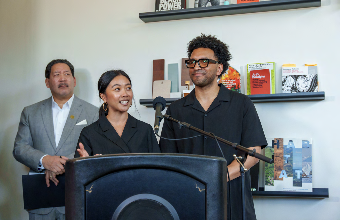 Three people speaking at a podium during La Union Studio's grand opening celebration, with a bookshelf of design objects behind them