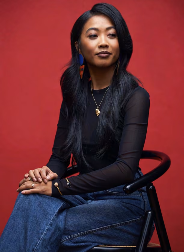 Woman with long dark hair seated against a red backdrop, wearing a black top and denim jeans