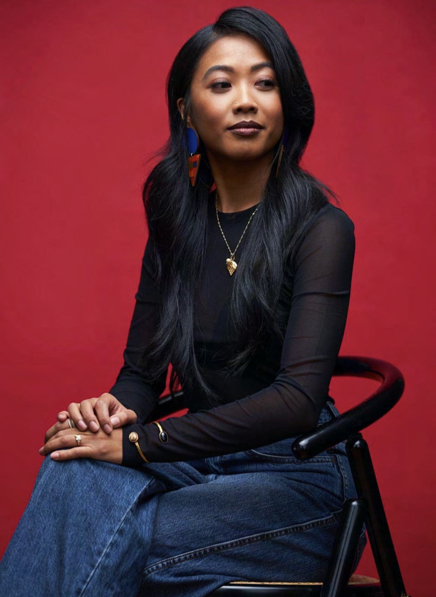 Woman with long dark hair seated against a red backdrop, wearing a black top and denim jeans
