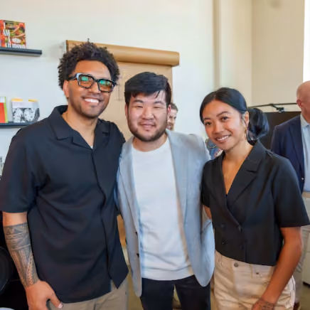 Three people smiling together indoors, including Sam Cho, at a community gathering