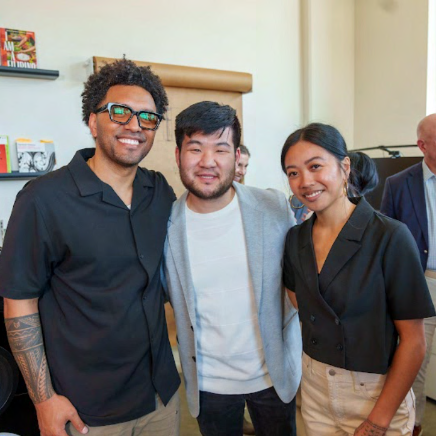 Three people smiling together indoors, including Sam Cho, at a community gathering