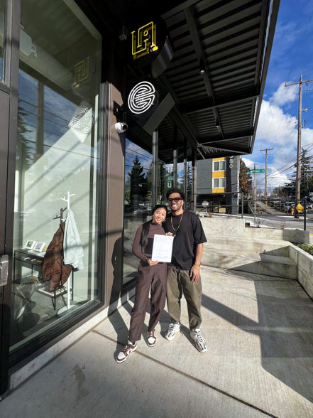 Two people standing outside a storefront holding a certificate, with skincare-related signage in the window