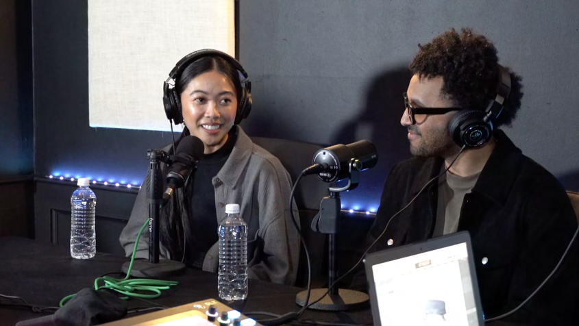 Woman and man sitting at a podcast recording setup wearing headphones, with microphones and water bottles on the table
