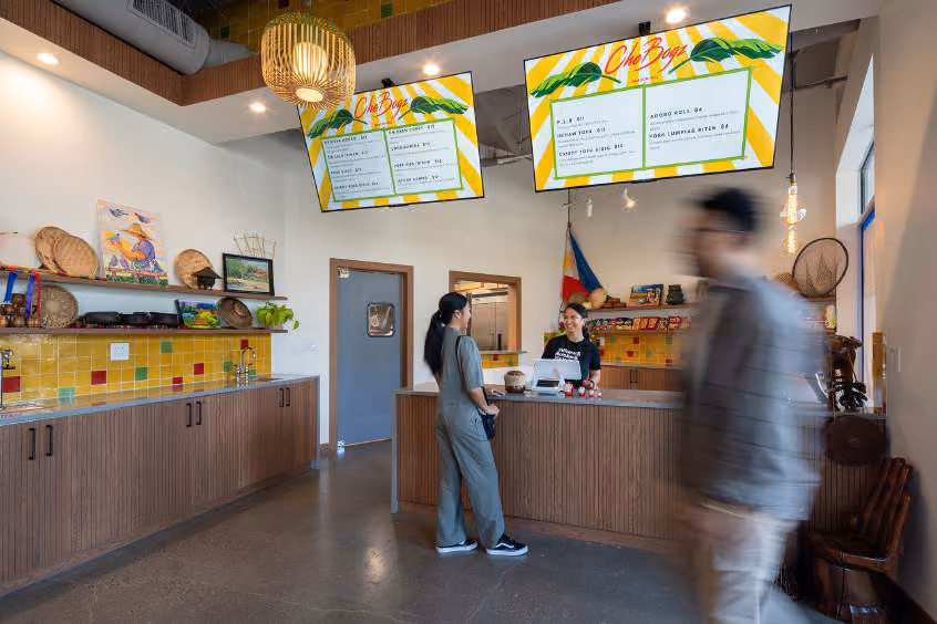 Restaurant counter with two hanging menu screens, a rattan pendant lamp, Philippine flag, and two people at the register while another walks past