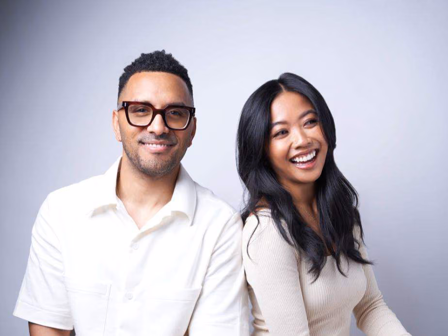 Man with glasses in a white shirt and woman with long dark hair in a cream top, smiling together against a gray backdrop