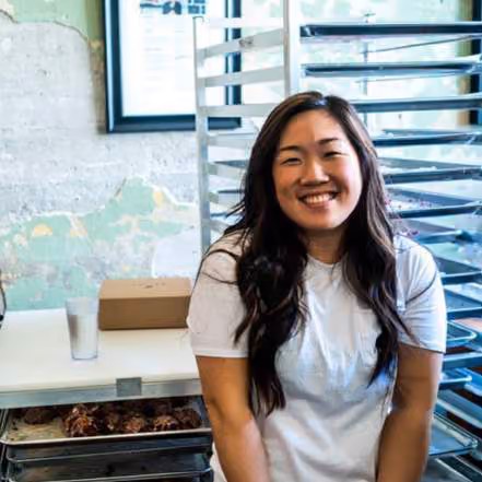 Woman with long dark hair smiling, standing in a doughnut shop with a metal shelving unit behind her
