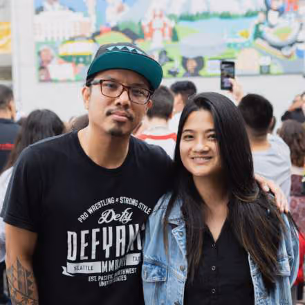 Man wearing a cap and graphic t-shirt standing with a woman in a denim jacket, in front of a colorful mural with a crowd nearby