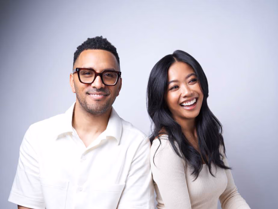 Man with glasses in a white shirt and woman with long dark hair in a cream top, smiling together against a gray backdrop