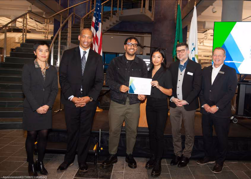 Six people posing together indoors, with one man and woman in the center holding a certificate, American and Washington state flags in the background