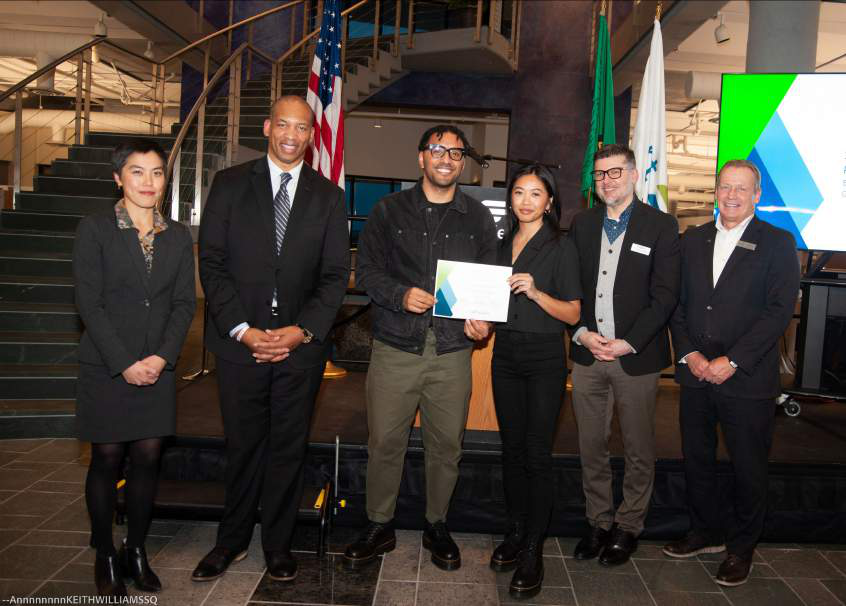Six people posing together indoors, with one man and woman in the center holding a certificate, American and Washington state flags in the background