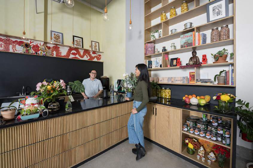 Interior with black countertops and wood cabinetry, a red dragon mural, and wall shelving stocked with jars, plants, and books; a man stands behind the counter and a woman stands in front