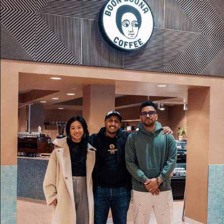 Three people standing together with arms around each other beneath a 'Boon Boona Coffee' sign at a cafe