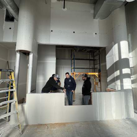 Three people talking inside a space under construction with exposed drywall, a ladder, and plastic sheeting over a counter