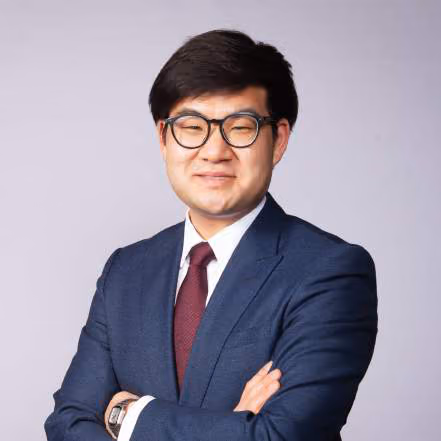 Man with glasses in a navy suit and red tie, arms crossed, against a lavender backdrop