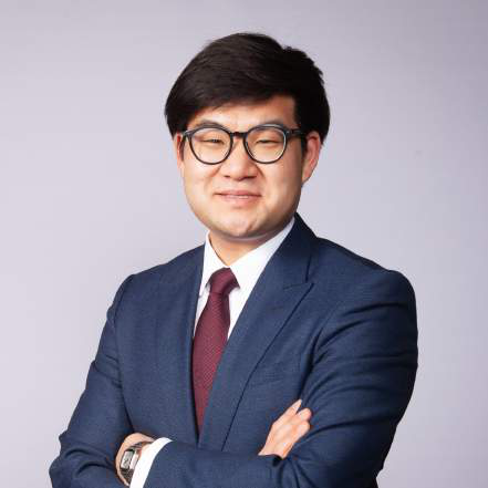 Man with glasses in a navy suit and red tie, arms crossed, against a lavender backdrop