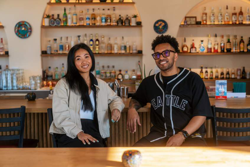 Woman with long dark hair in a cream jacket and man in a black 'Seattle' baseball jersey, seated at a bar with shelves of bottles behind them