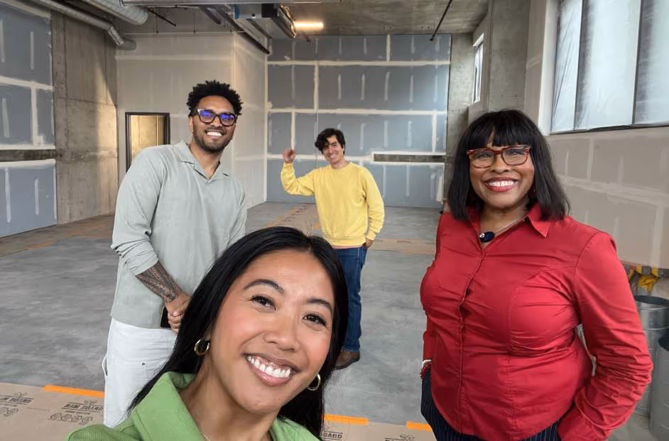 Four people posing in an unfinished construction space, one smiling closely at the camera, others standing behind