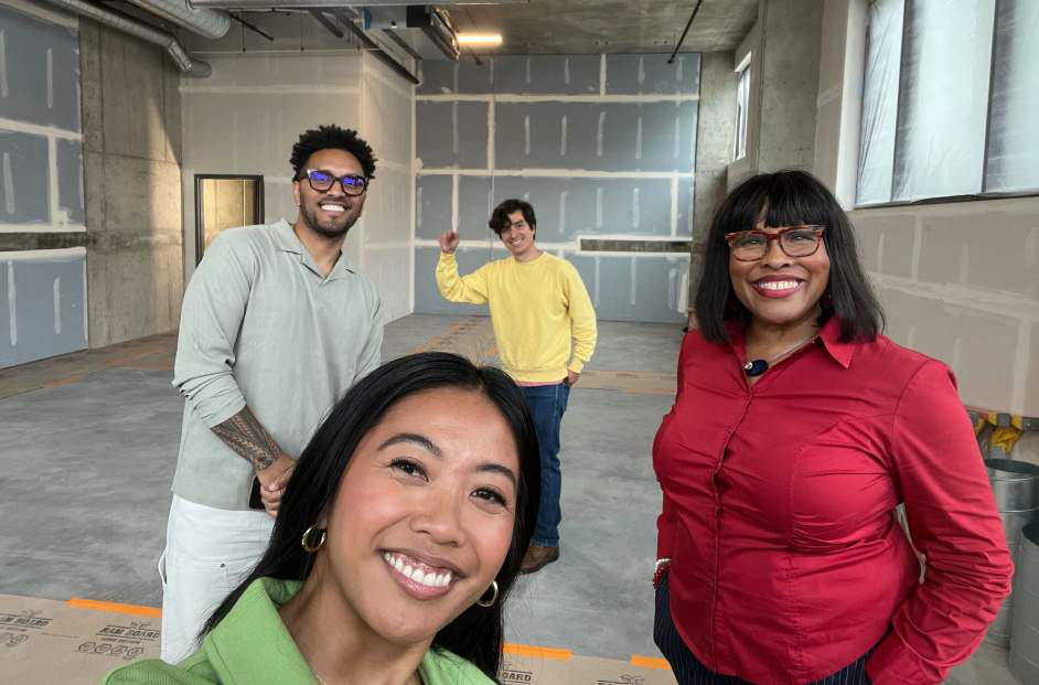 Four people posing in an unfinished construction space, one smiling closely at the camera, others standing behind