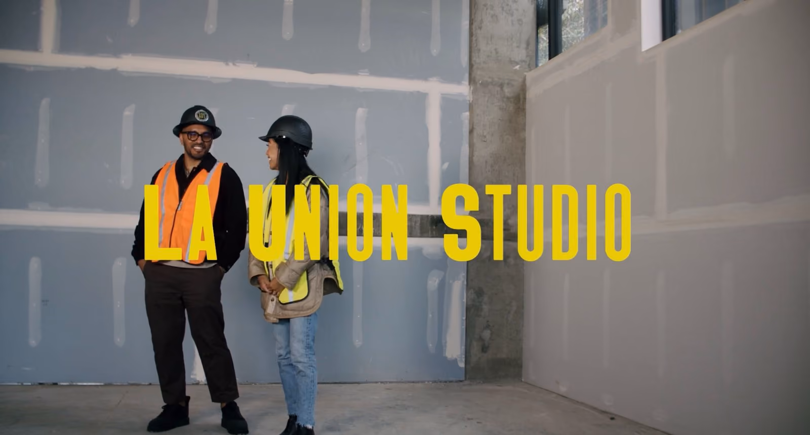 Two people in hard hats and safety vests standing in an unfinished space, with large yellow "La Union Studio" text overlaid across the image