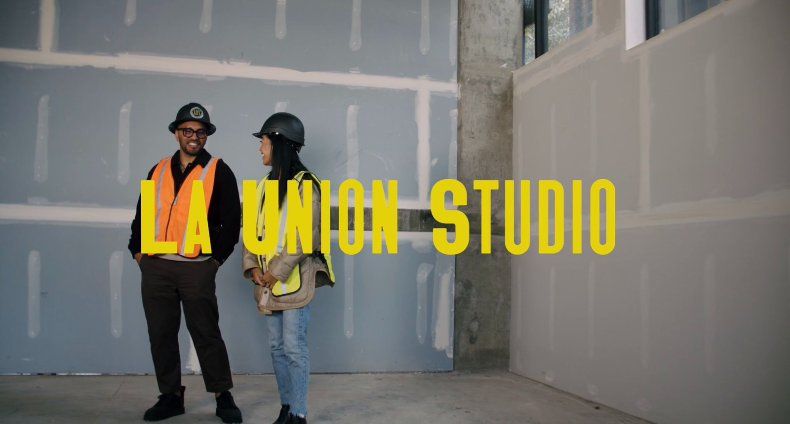 Two people in hard hats and safety vests standing in an unfinished space, with large yellow "La Union Studio" text overlaid across the image