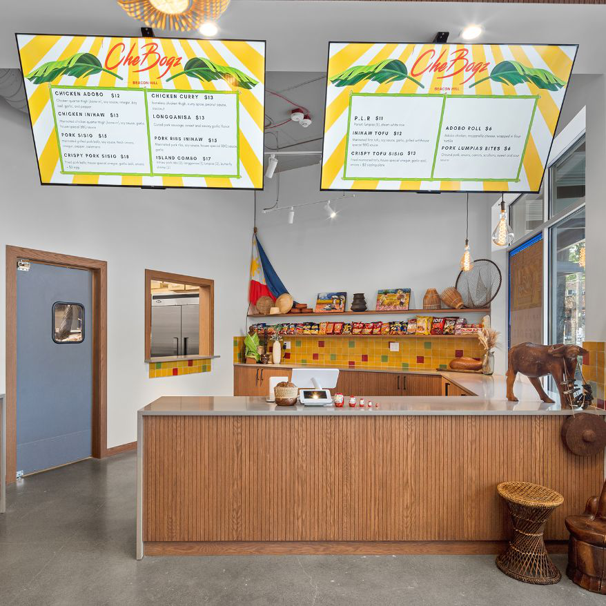 CheBogz Filipino Food interior with two hanging menu screens, wood counter, Philippine flag, and yellow tile backsplash