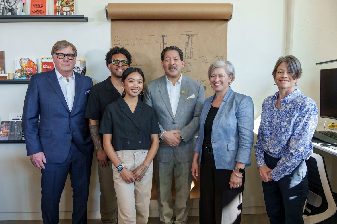 Six people standing shoulder to shoulder inside the La Union Studio office, in front of a wall pinned with a large architectural drawing and shelves of books and objects.
