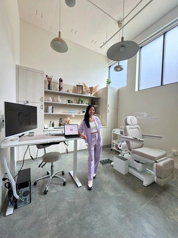 Sonia-Lynn Abenojar standing in the Emblem Skincare treatment room, beside a desk and a reclining treatment chair, with shelves of skincare products and pendant lamps overhead.