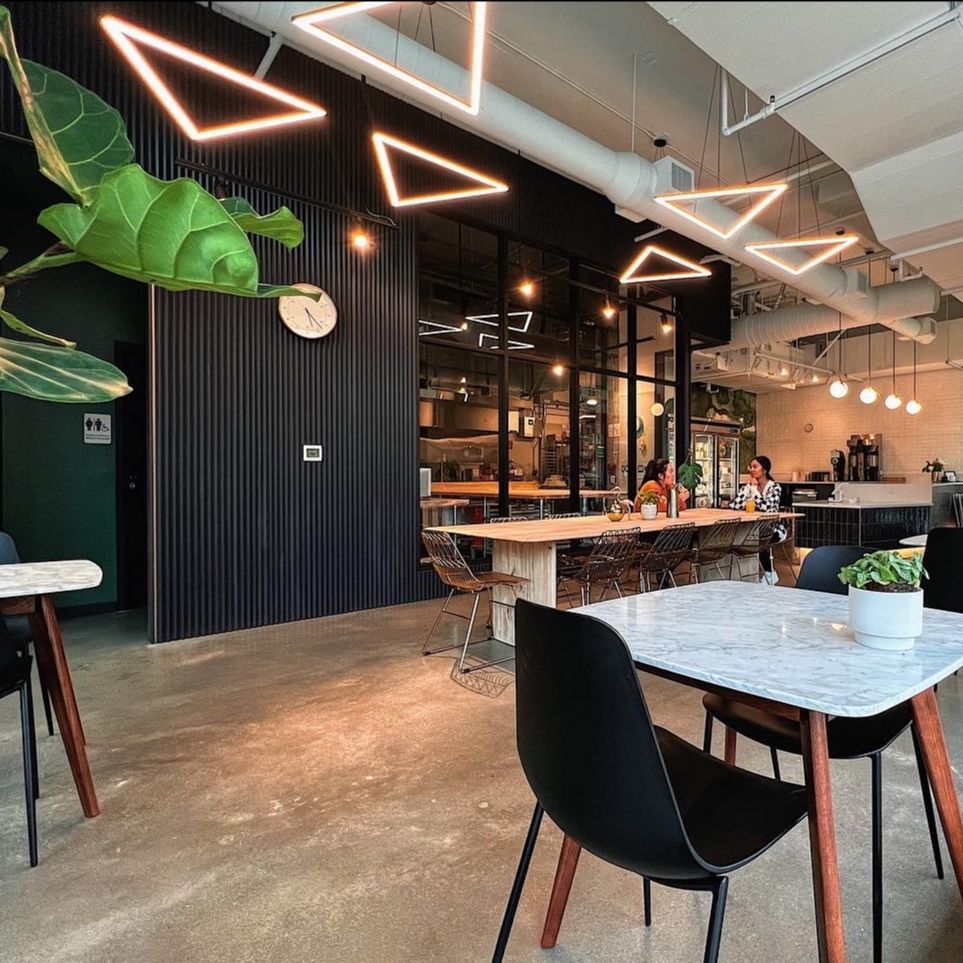 The seating area at Raised Doughnuts & Cakes, with angular neon triangle light fixtures suspended from the ceiling, dark slatted walls, marble-topped tables, black chairs, and a hanging plant.
