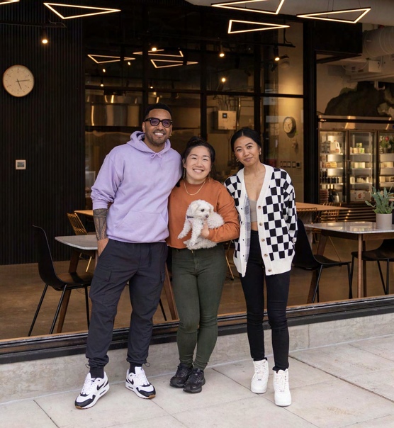 Three people standing in front of the Raised Doughnuts & Cakes storefront window at night, one of them holding a small white dog.