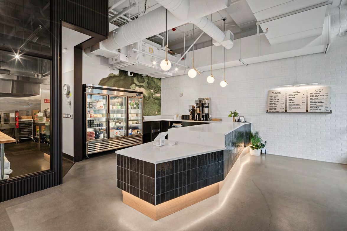The service counter at Raised Doughnuts & Cakes: a curved dark-tiled counter with a white stone top and illuminated base, a glass pastry case, exposed white ductwork, white brick walls, and menu boards.