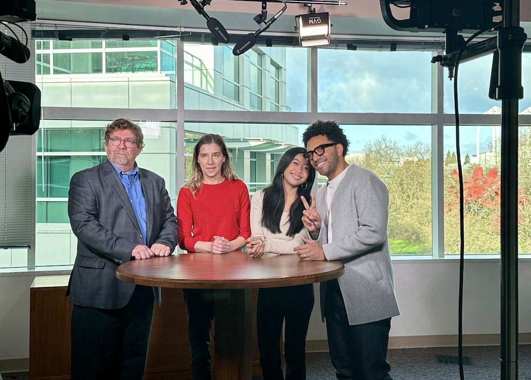 Four people standing around a round table in a television studio, with lighting rigs overhead and windows looking onto trees behind them.