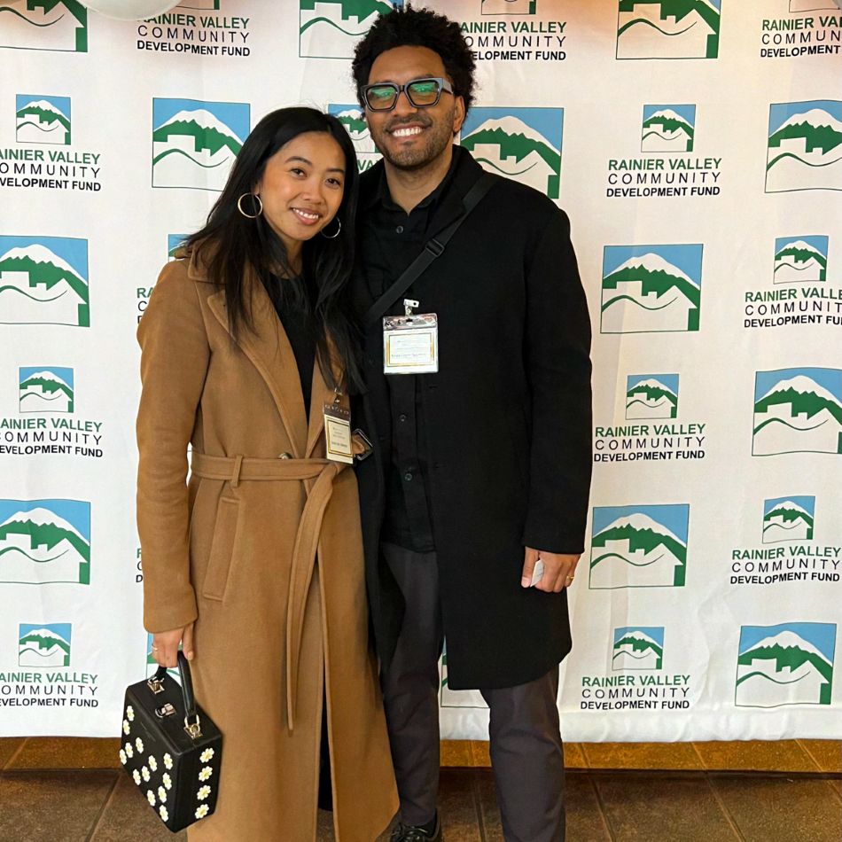 Two people posing side by side in front of a step-and-repeat banner printed with the Rainier Valley Community Development Fund logo.