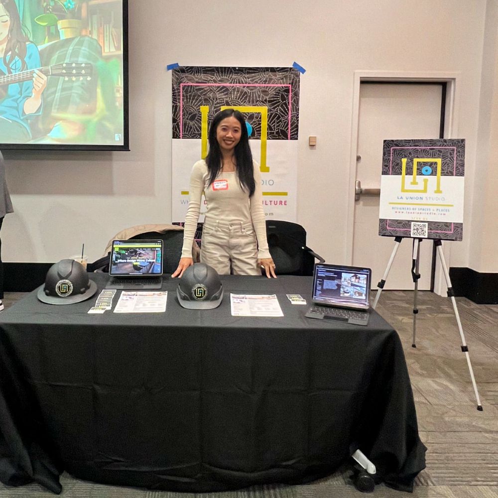A person standing behind a black-draped exhibition table set with two laptops, printed handouts, and two branded hard hats, with a La Union Studio banner and easel poster behind them.