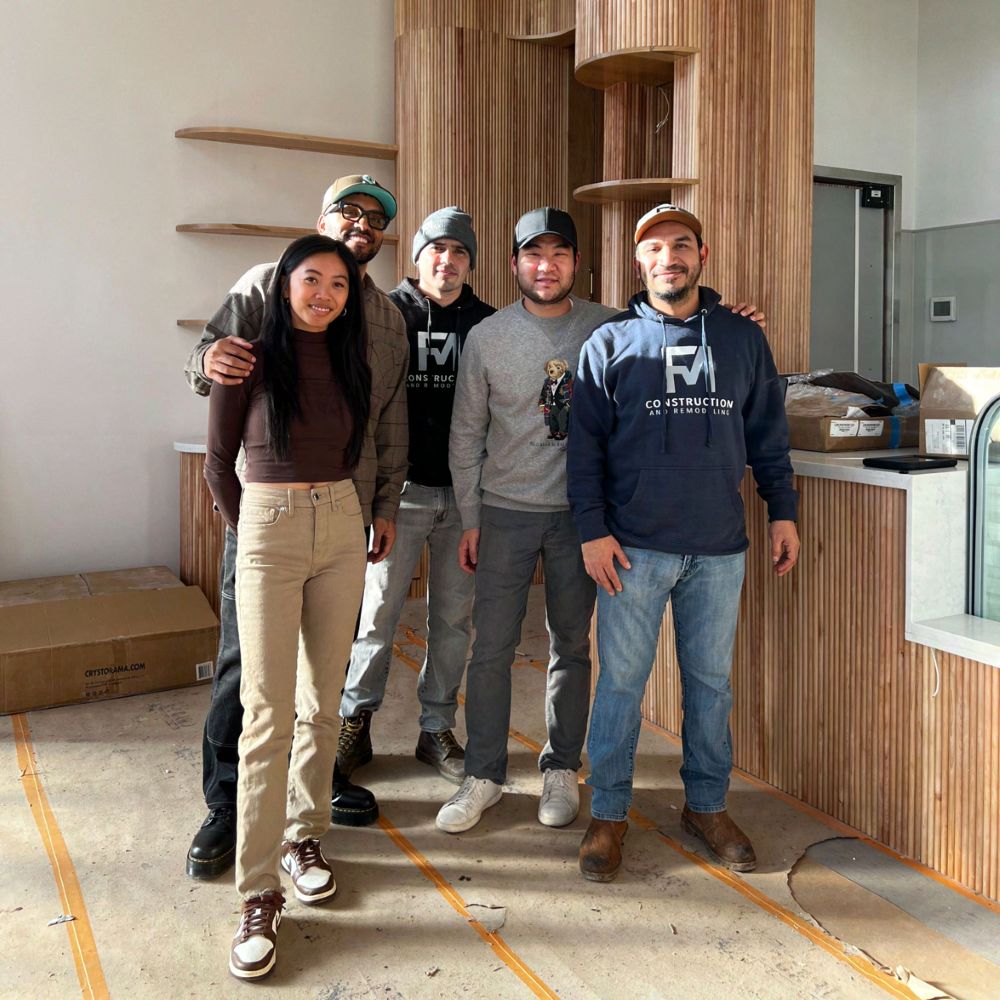 Five people standing shoulder to shoulder inside the unfinished La Union Studio HQ, in front of a curved wall of vertical wood slats and floating shelves, with bare concrete floors marked in orange tape, cardboard boxes, and a partly built service counter behind them.