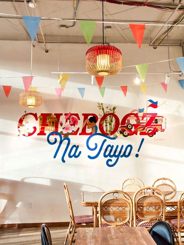The dining room at CheBogz Filipino Food, with a large mural in red and blue script reading "CheBogz Na Tayo!" alongside a decorated jeepney, colorful triangular bunting and woven pendant lanterns strung across the ceiling, and rattan chairs at a wooden table.
