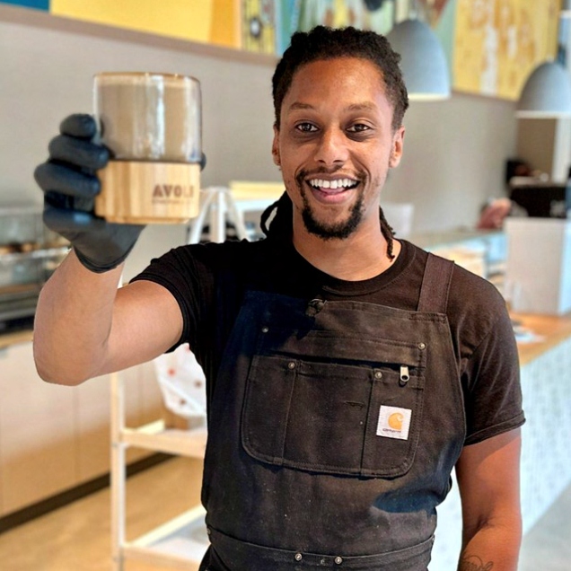 Solomon Dubie smiling and holding a cup of coffee up toward the camera inside Cafe Avole, wearing a dark work apron over a t-shirt.