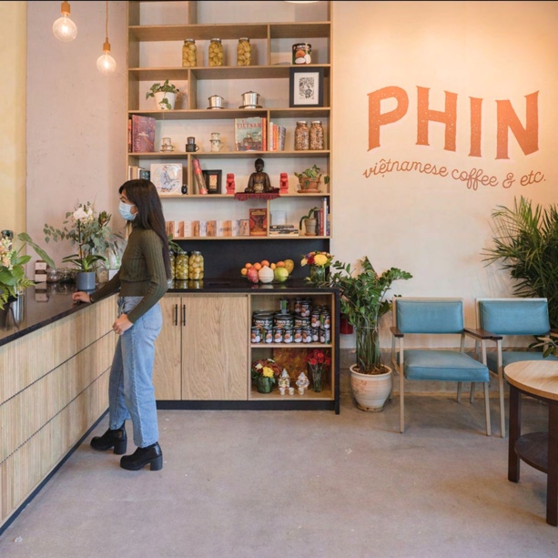 The interior of Phin Vietnamese Coffee, with an orange hand-painted wall sign reading "Phin — Vietnamese coffee & etc," light wood shelving stocked with jars and packaged goods, teal upholstered chairs, potted plants, and a person browsing the shelves.