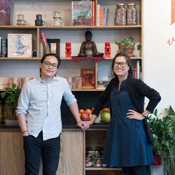 Two people standing side by side in front of the shelving at Phin Vietnamese Coffee, which holds books, a Buddha figure, bags of coffee, and a bowl of fruit on the counter below.