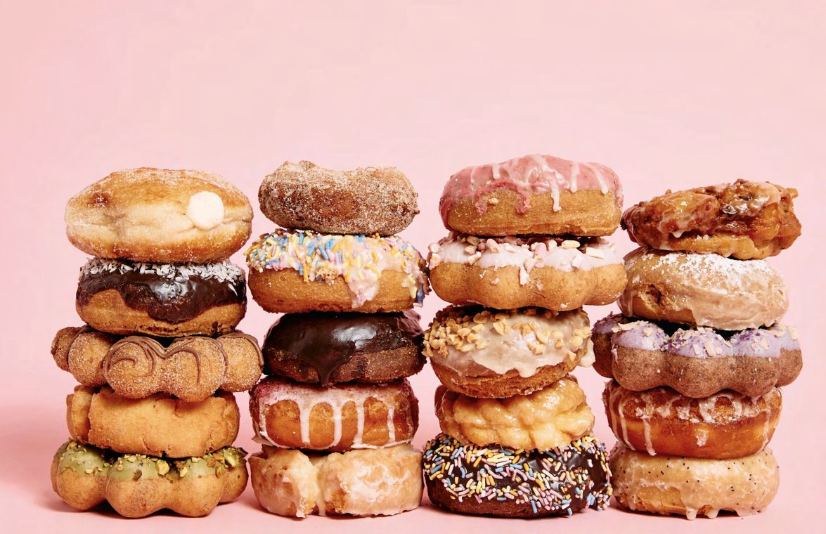 Four tall stacks of assorted doughnuts — glazed, chocolate-frosted, sprinkled, sugared, and filled — arranged against a plain pink background.