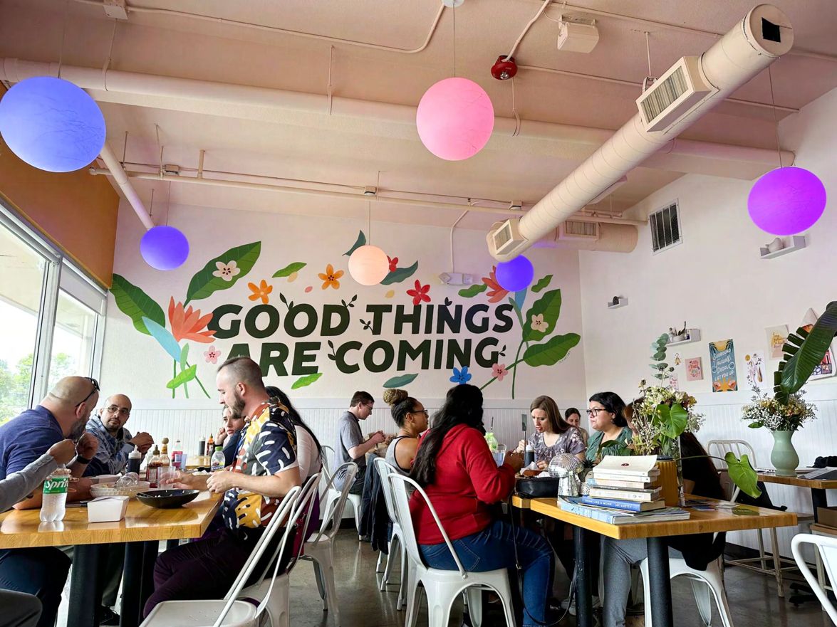 Diners seated at wooden tables inside Ta Joia, under a wall mural of colorful flowers and foliage reading "Good Things Are Coming," with spherical pendant lights in blue, pink, and purple hanging from the ceiling and white metal chairs throughout.