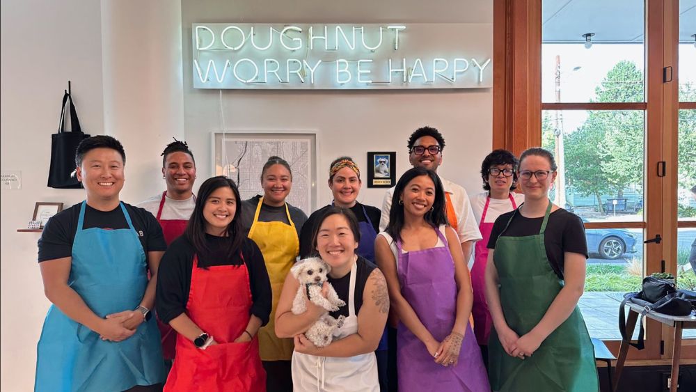 About ten members of the La Union Studio team standing together in brightly colored aprons — blue, red, yellow, purple, green — under a pale blue neon sign reading "Doughnut worry be happy," with one person holding a small white fluffy dog and daylight coming through the windows behind them.