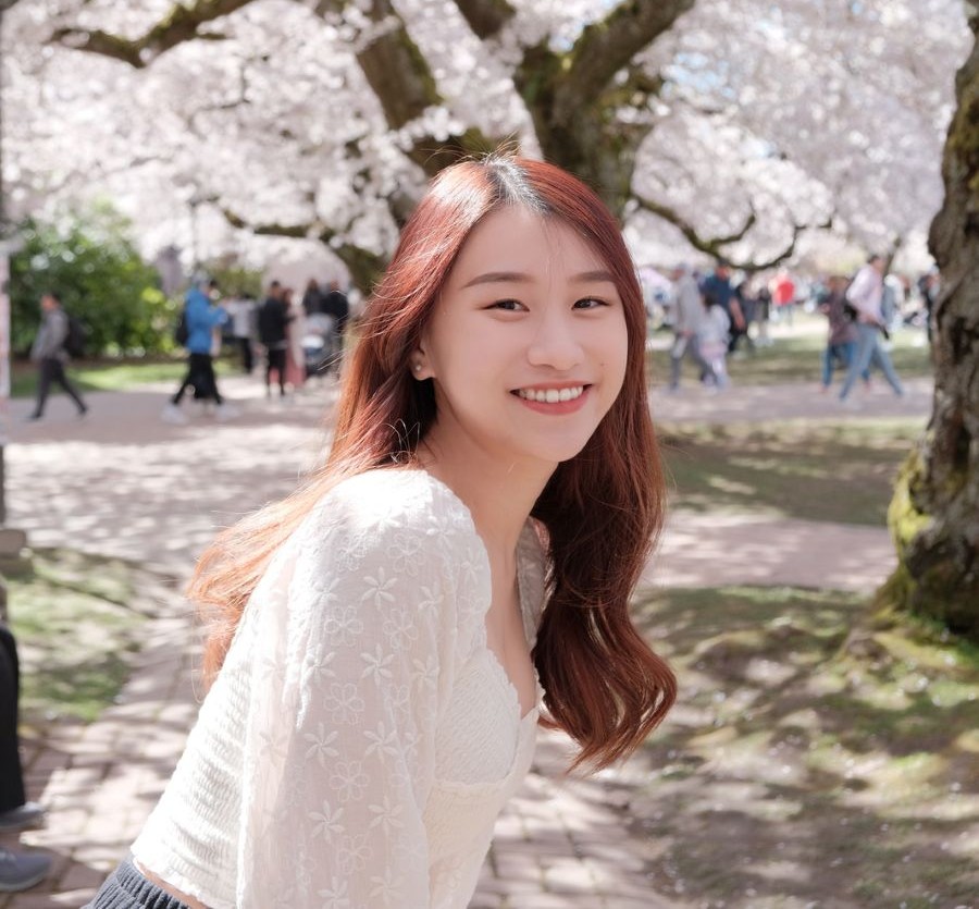 Aure Yan, the studio's new community design intern, smiling over her shoulder in a white lace top while standing on a path beneath cherry trees in full pink blossom, with people walking in the background.
