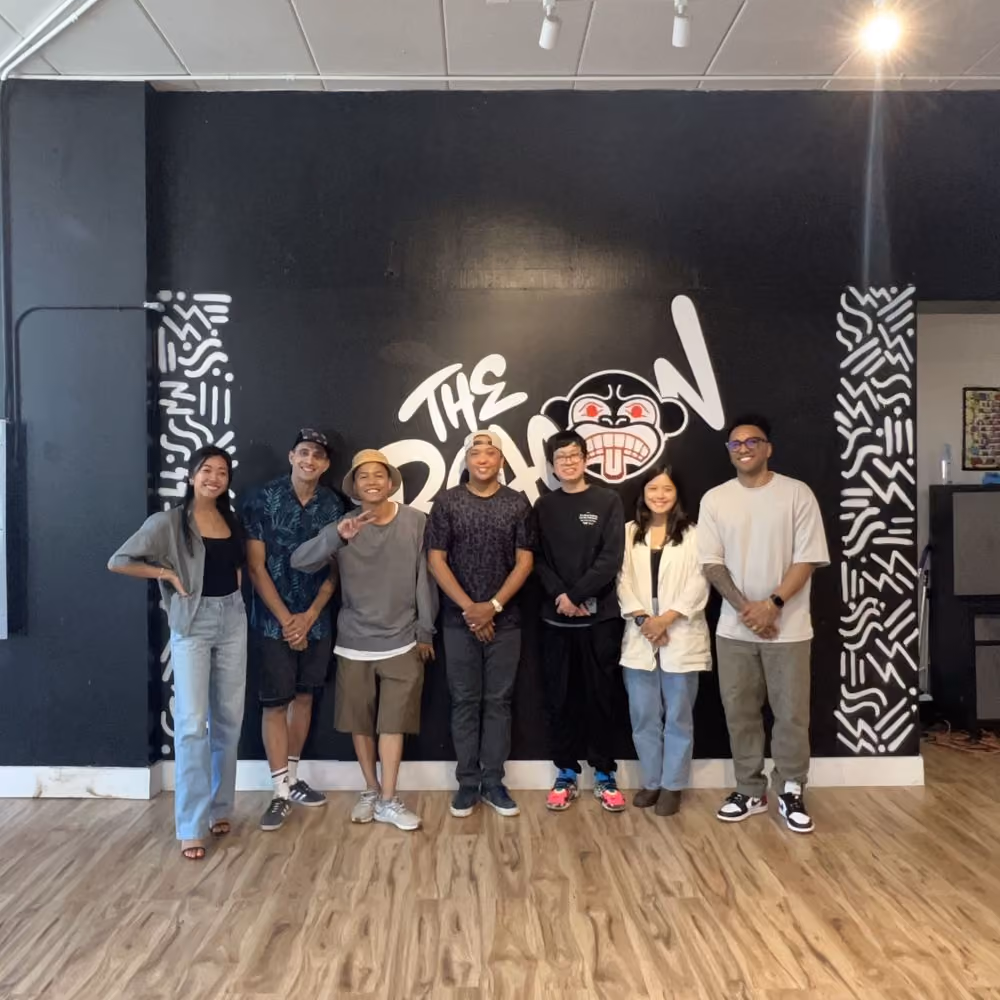 Seven people standing in a row on a light wood floor in front of a dark wall painted with a large graffiti-style mural reading "The Beacon," flanked by black-and-white geometric patterning.